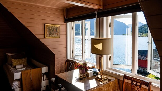 A cozy cabin room with a wooden interior, a dining table with chairs, a lamp, and a view of a lake and mountains through large windows. - Fjærland Fjordstove Hotell (Sogndal)