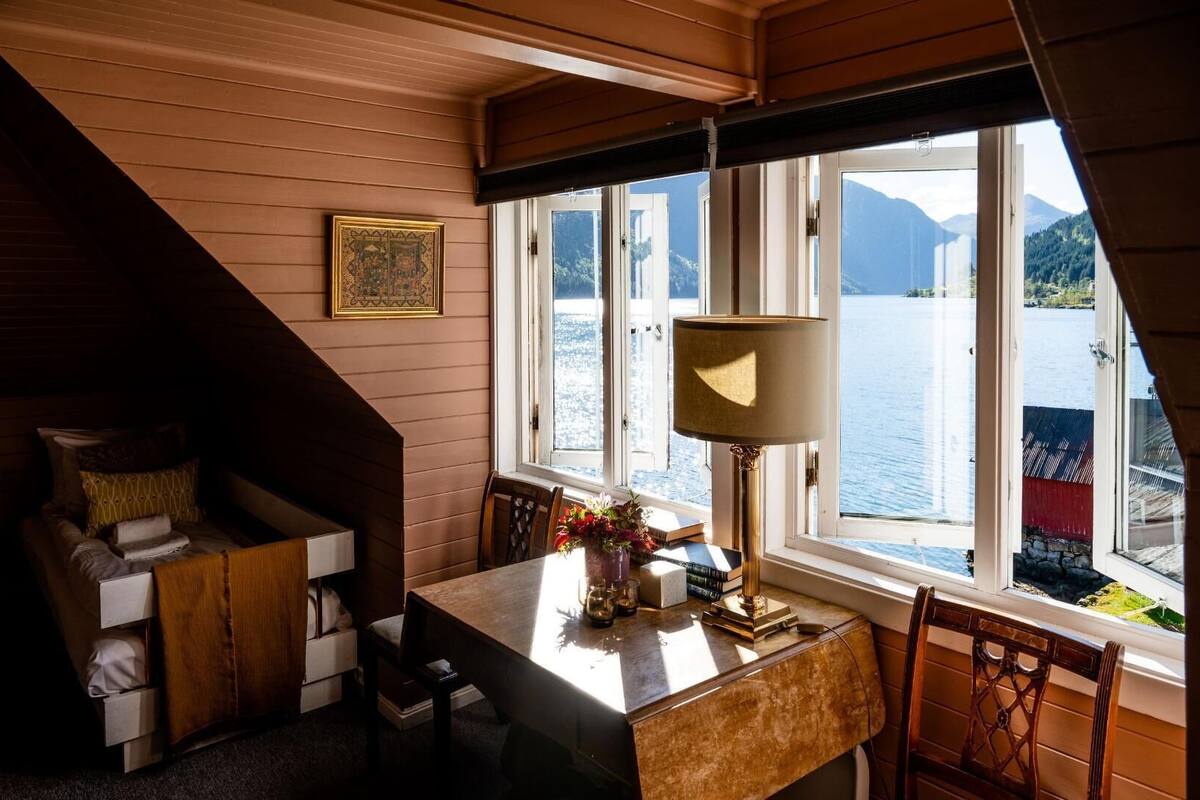 Ein gemütliches Blockhausszimmer mit Holzvertäfelung, einem Esstisch mit Stühlen, einer Lampe und Blick auf einen See und Berge durch große Fenster.