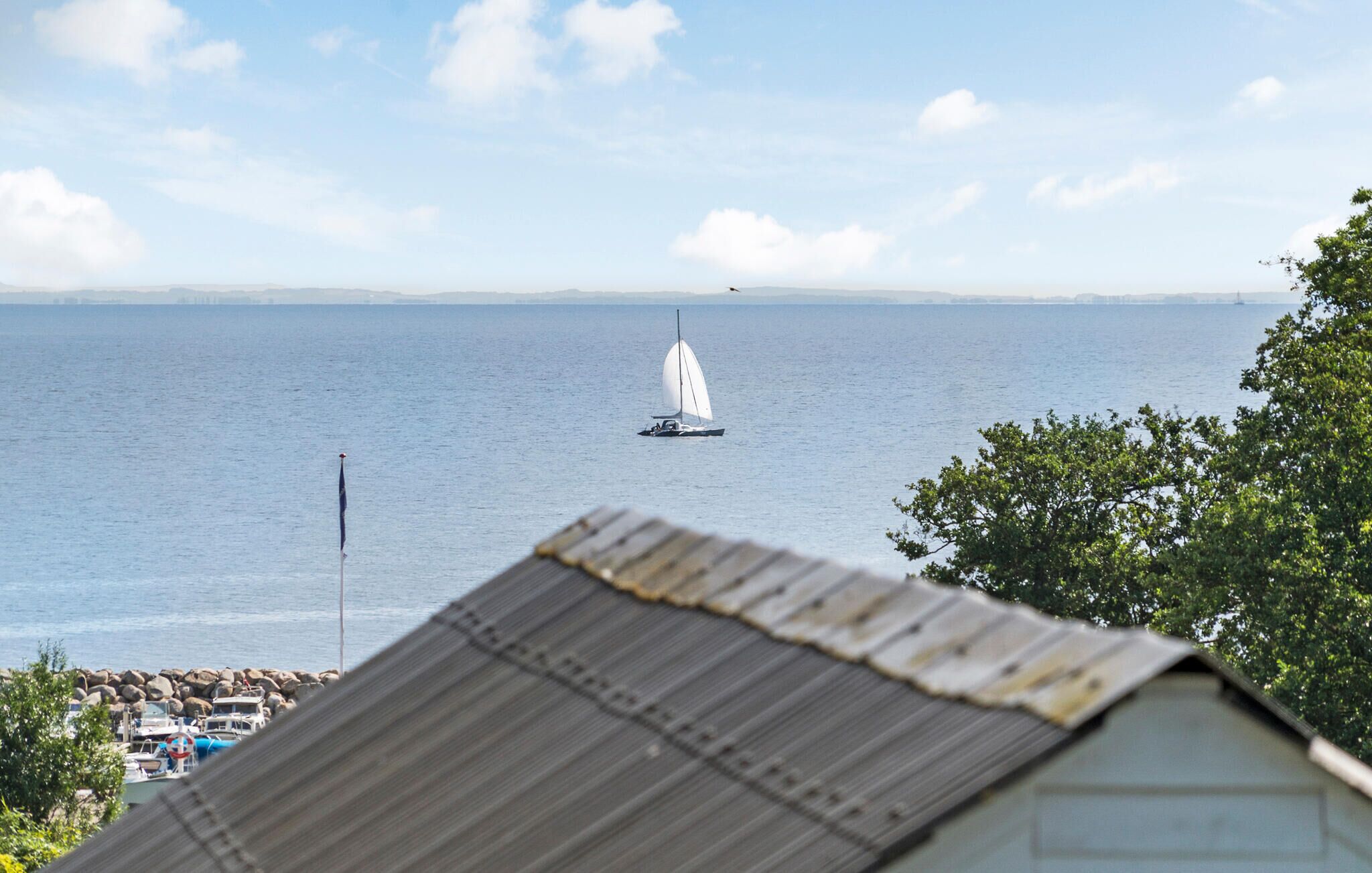 A sailboat on a calm sea with a distant shoreline and a building with a corrugated roof in the foreground.