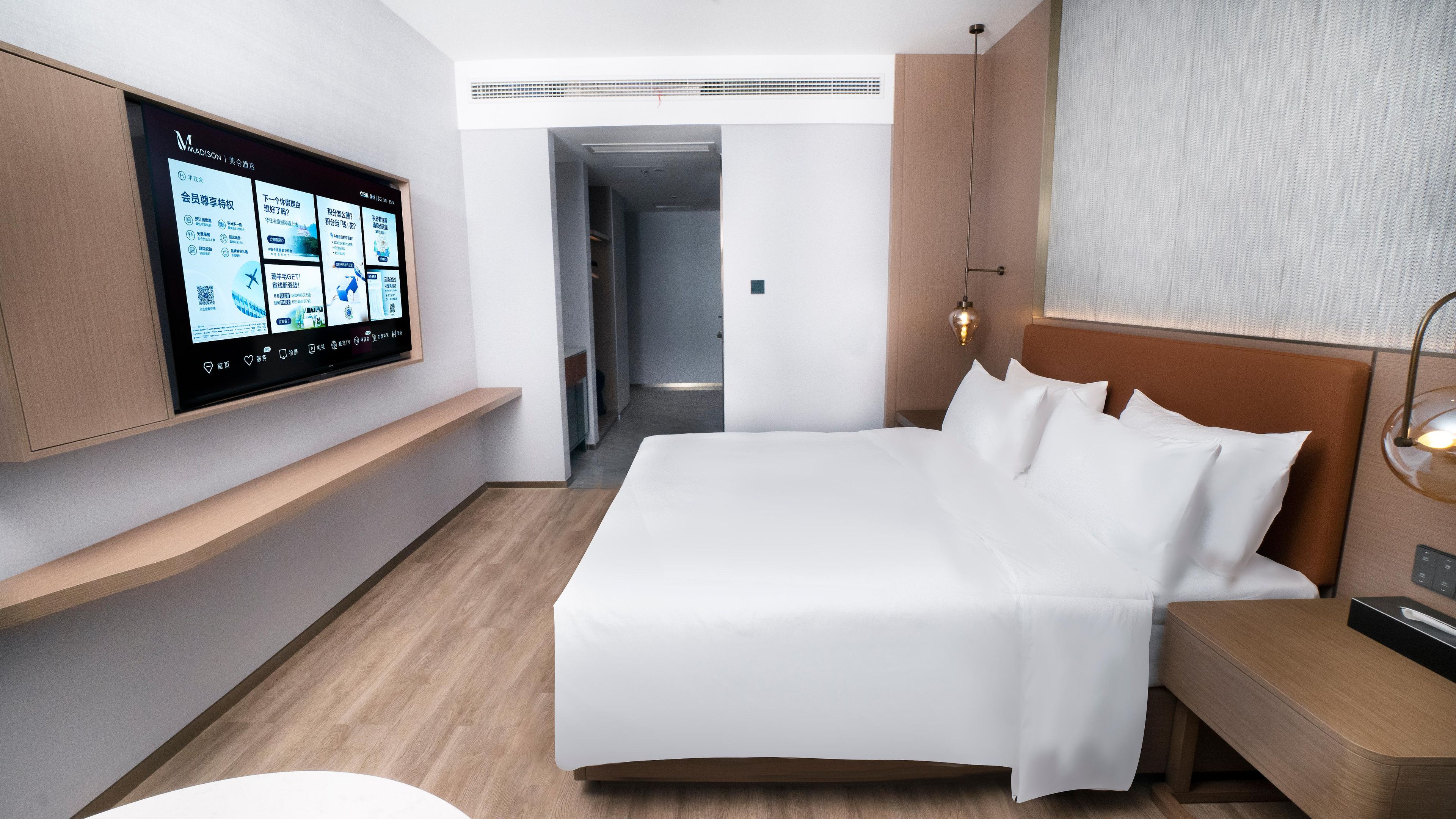 A modern hotel room with a large bed, a mounted TV displaying content, a bedside table, and a nightstand.