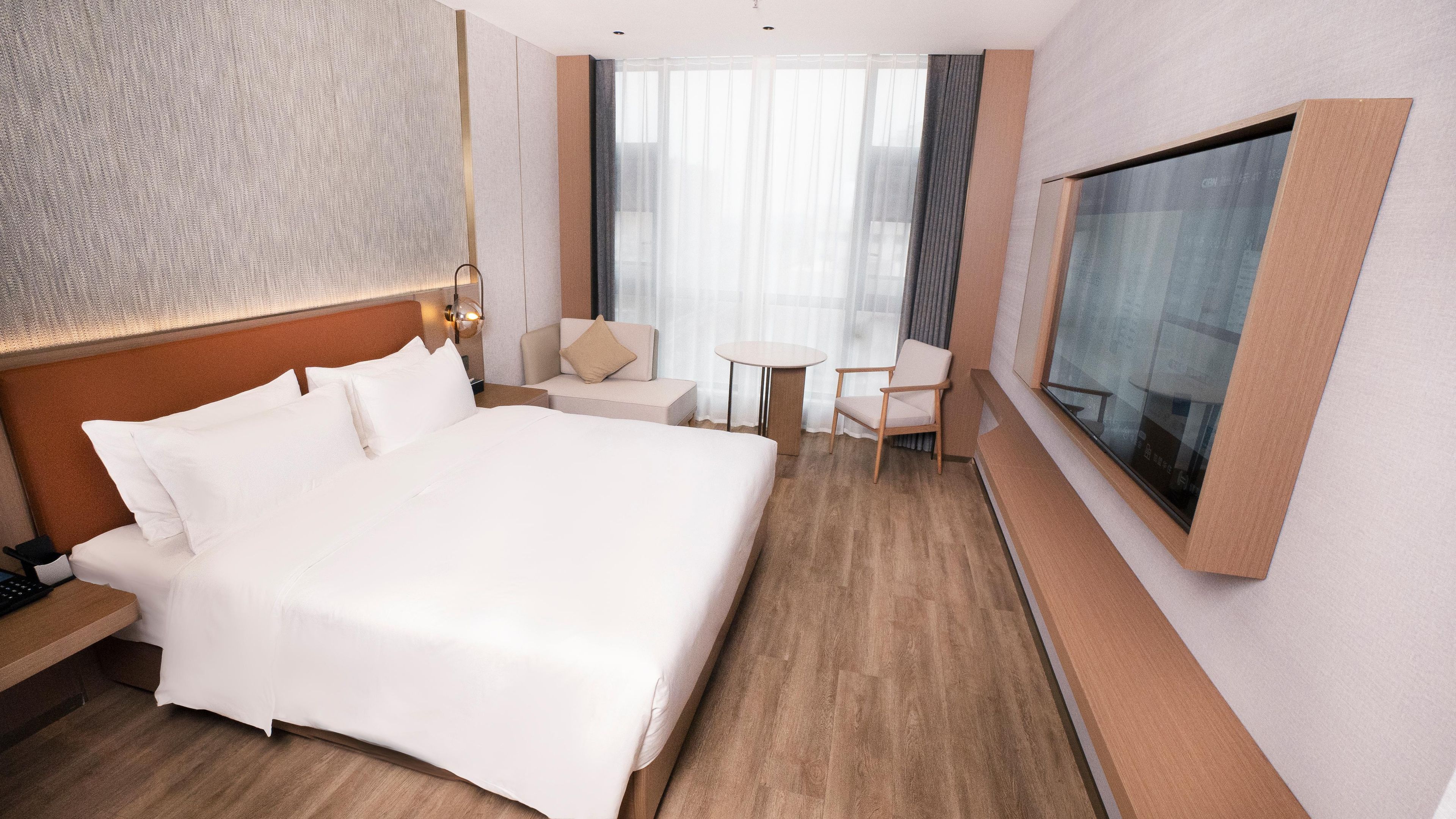 A hotel room with a large bed, a desk, a chair, a small table, and a TV mounted on the wall.