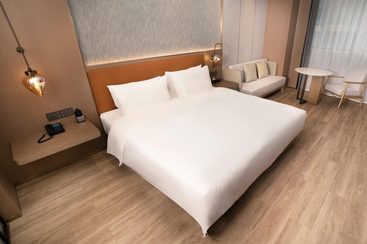 A hotel room with a large bed, bedside tables, a sofa, a small table, and a wall-mounted lamp.