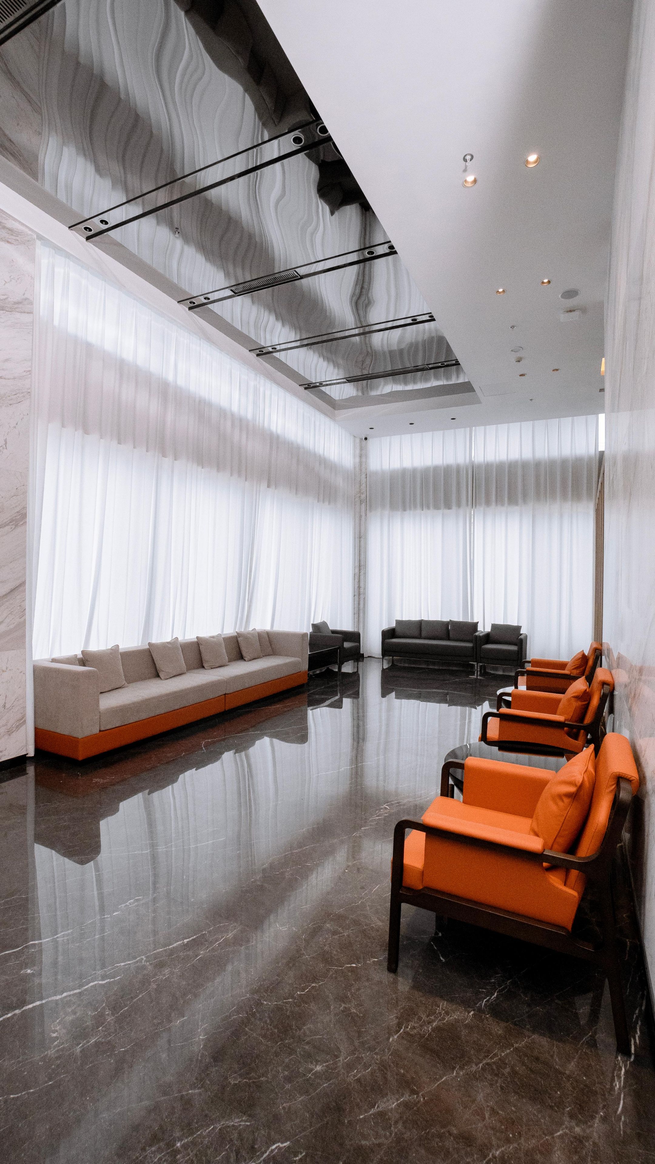 A spacious, modern waiting area with a marble floor, a long sofa, and several orange chairs.
