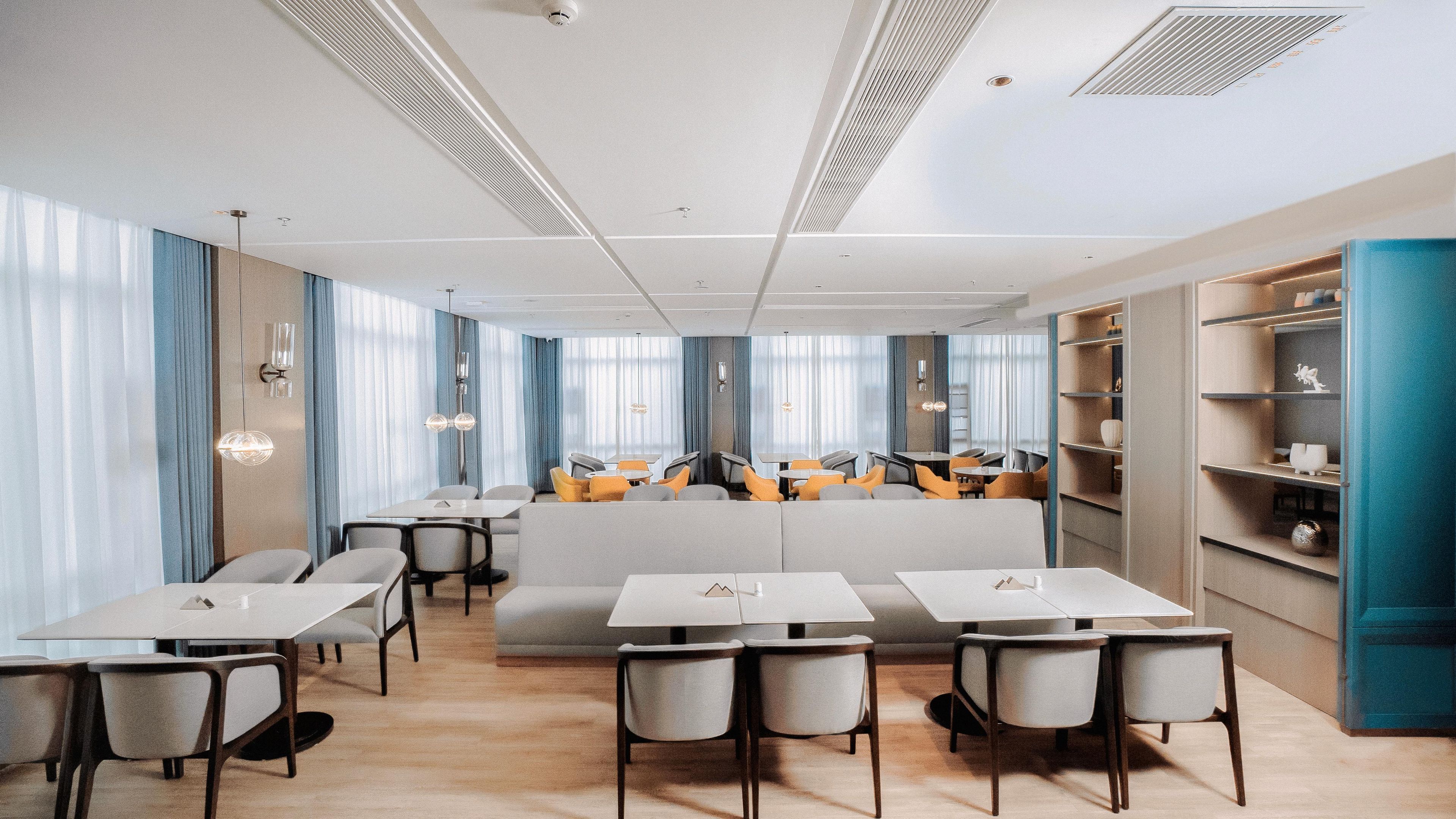 A modern dining area with tables and chairs, a central seating area, and a bookshelf.