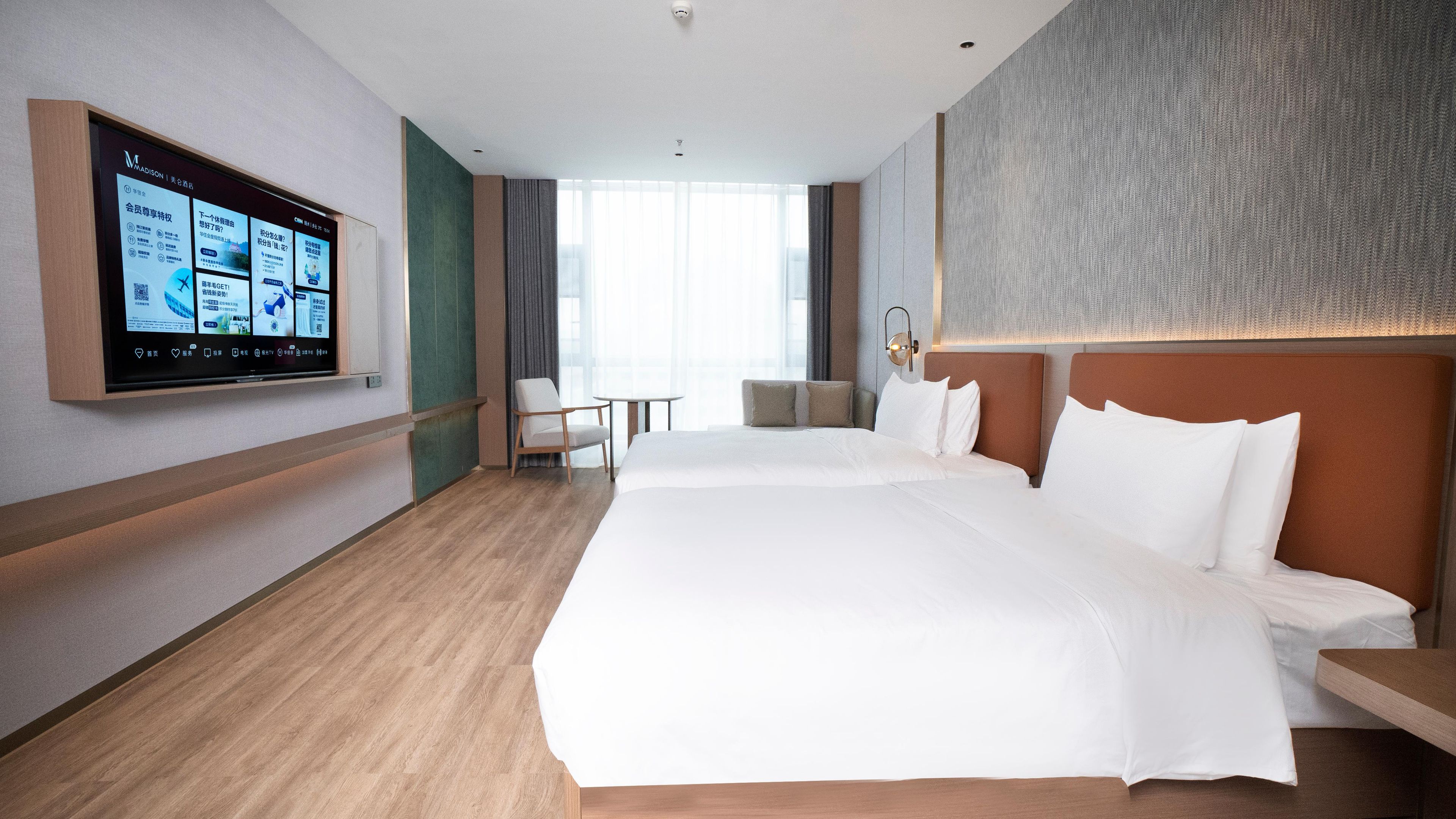 Ett hotellrum med en stor säng, ett skrivbord, en stol och en tv som är monterad på väggen och visar information.