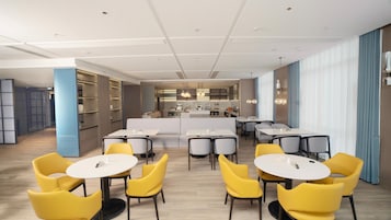 Un área de comedor moderna con sillas amarillas y mesas blancas, una cocina al fondo y una cortina azul a la derecha.