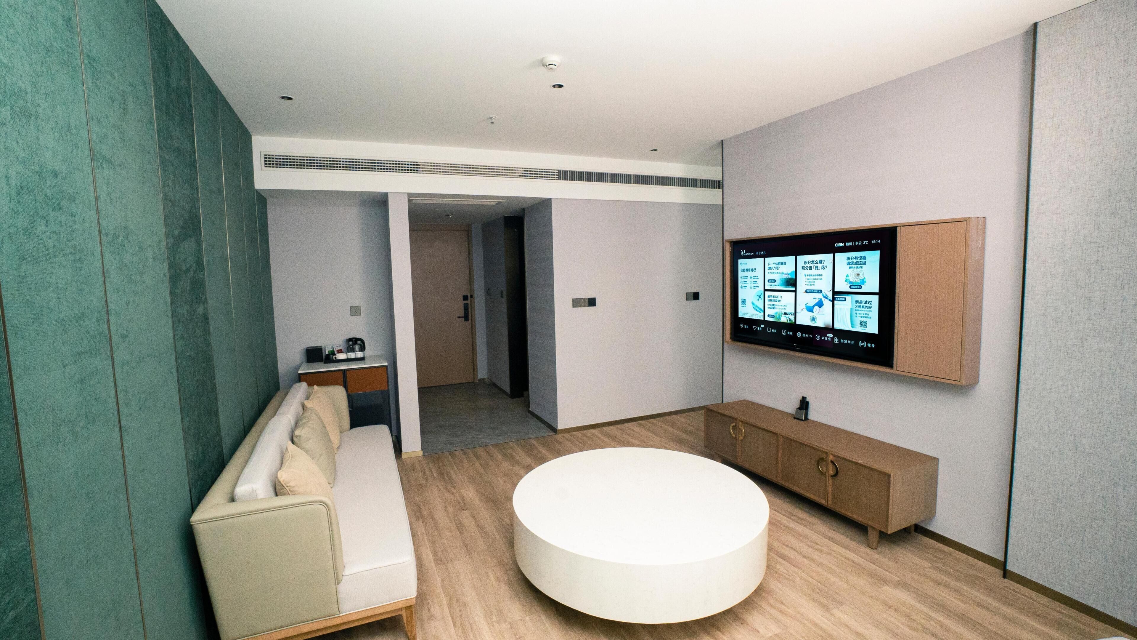 A modern bathroom with a round bathtub, a bench, a vanity with a mirror, and a wall-mounted TV displaying information.