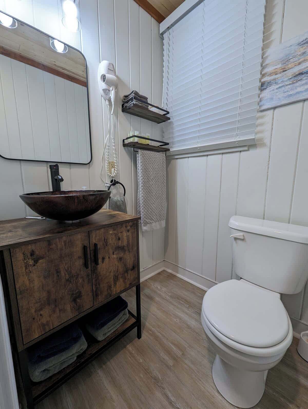 A bathroom with a wooden vanity, a large mirror, a white toilet, and a window with blinds.