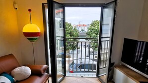 A living room with a brown leather sofa, a glass coffee table, a TV on a wooden stand, and a balcony with a view of the street and buildings.