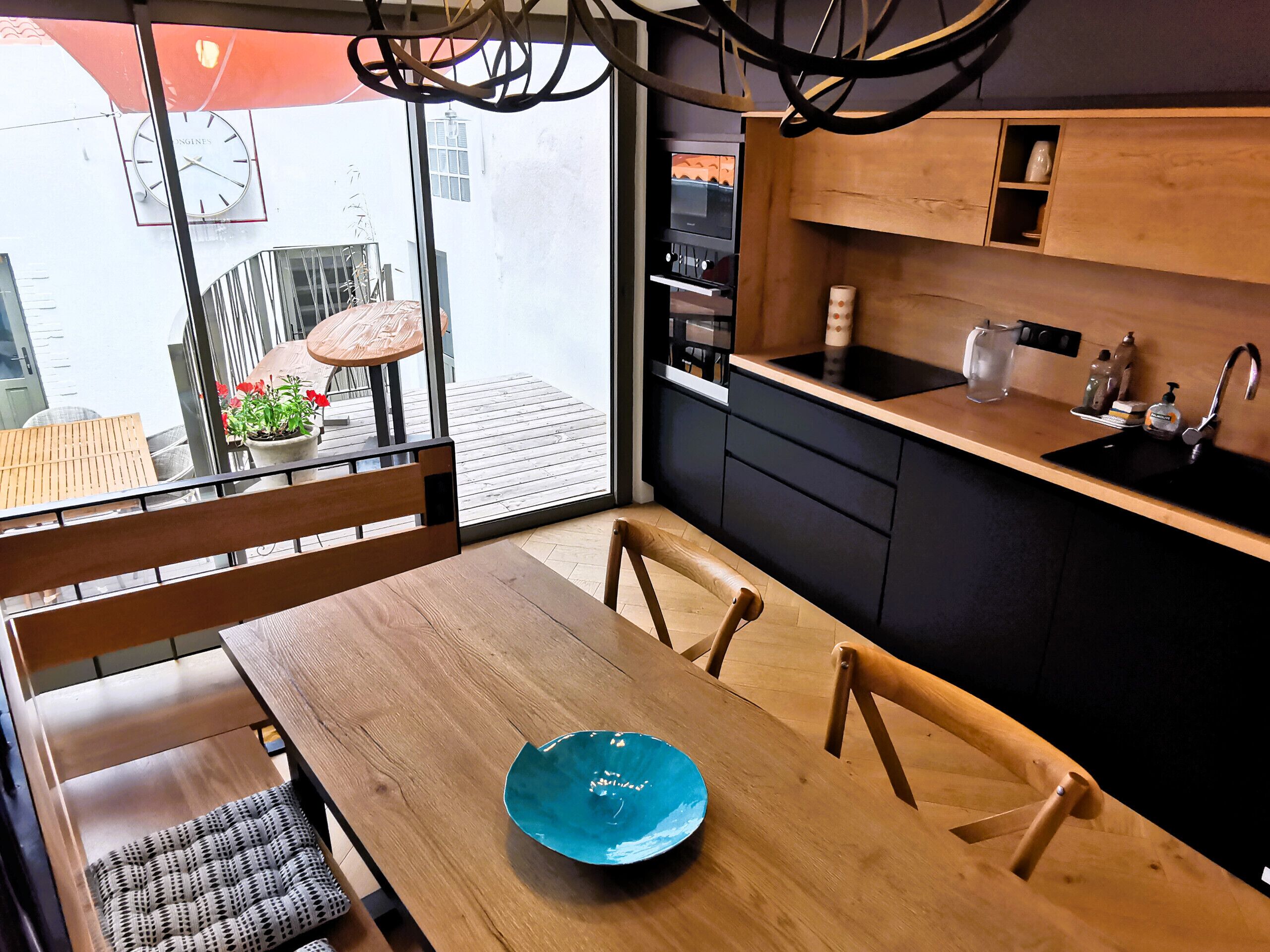 A modern kitchen with a black countertop, wooden cabinets, and a built-in oven. There is a dining area with a wooden table and chairs, and a blue bowl on the table.