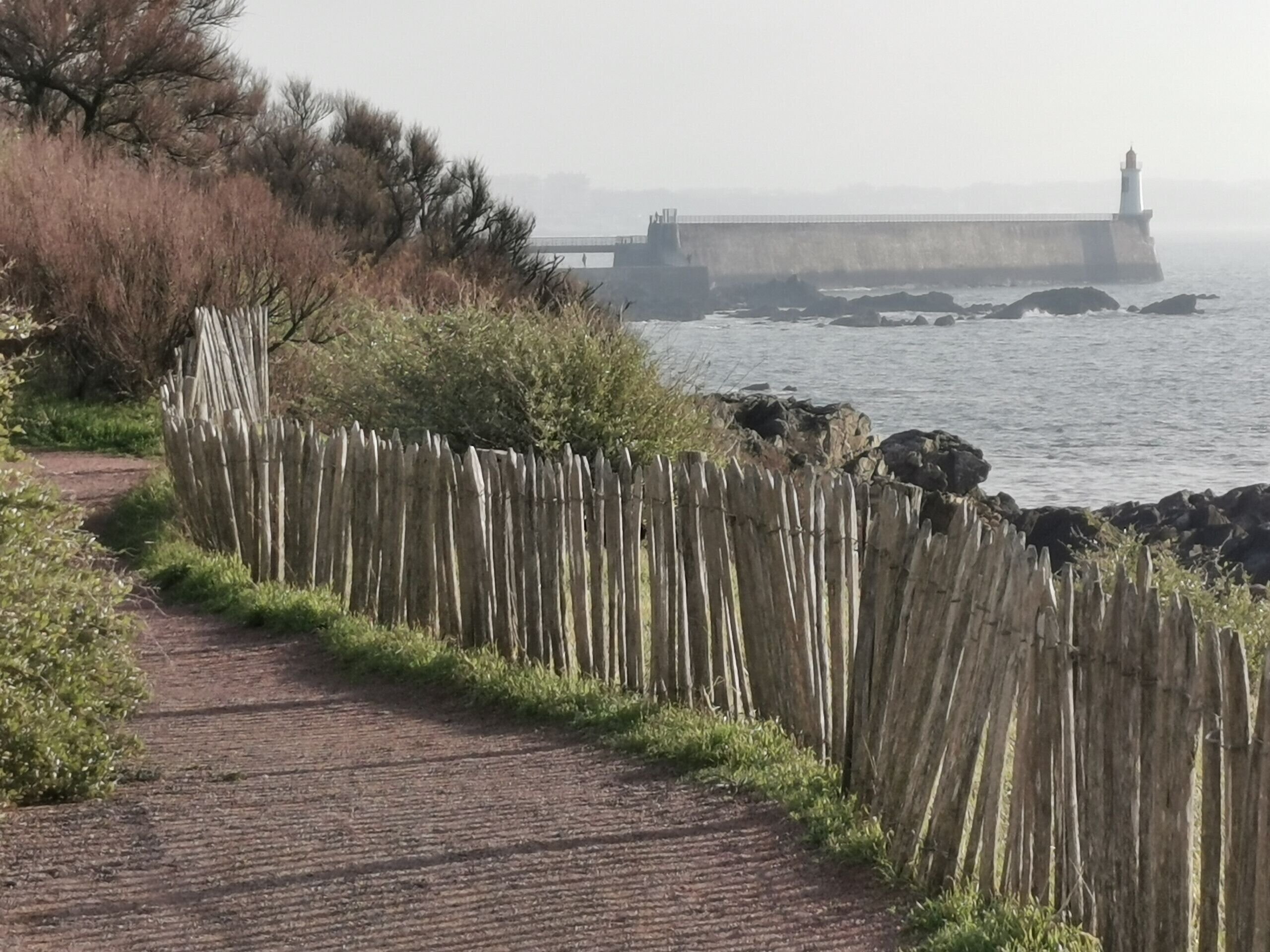 A coastal path with a wooden fence, a brick walkway, and a lighthouse in the distance.