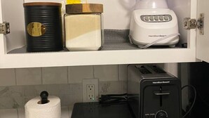 Una cucina con un mobiletto bianco che contiene un frullatore, un contenitore nero e un oggetto giallo. Sotto, si trova un forno a microonde e un tostapane.