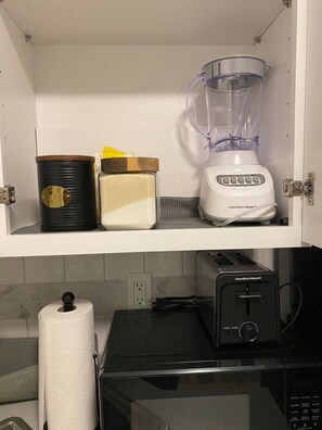 Une cuisine avec un meuble blanc contenant un mixeur, un récipient noir et un objet jaune. En dessous, on trouve un four à micro-ondes et un grille-pain.