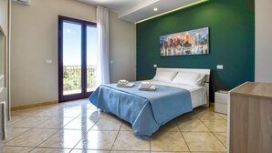 A hotel room with a bed, a painting on the wall, a balcony with a view, and a desk with a chair.