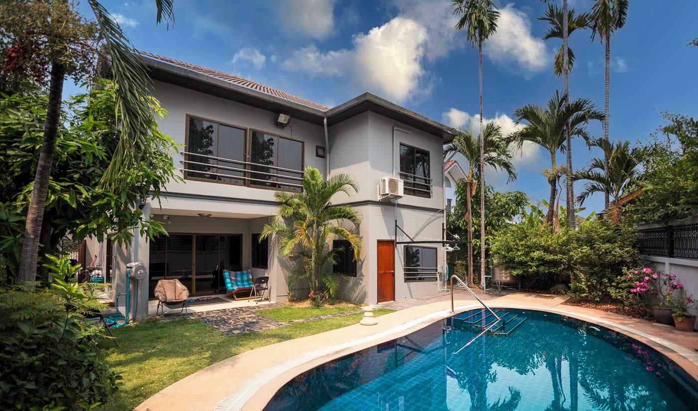 A two-story house with a swimming pool, surrounded by lush greenery and palm trees.