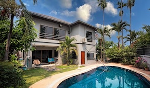 Une maison à deux étages avec une piscine, entourée d’une végétation luxuriante et de palmiers.