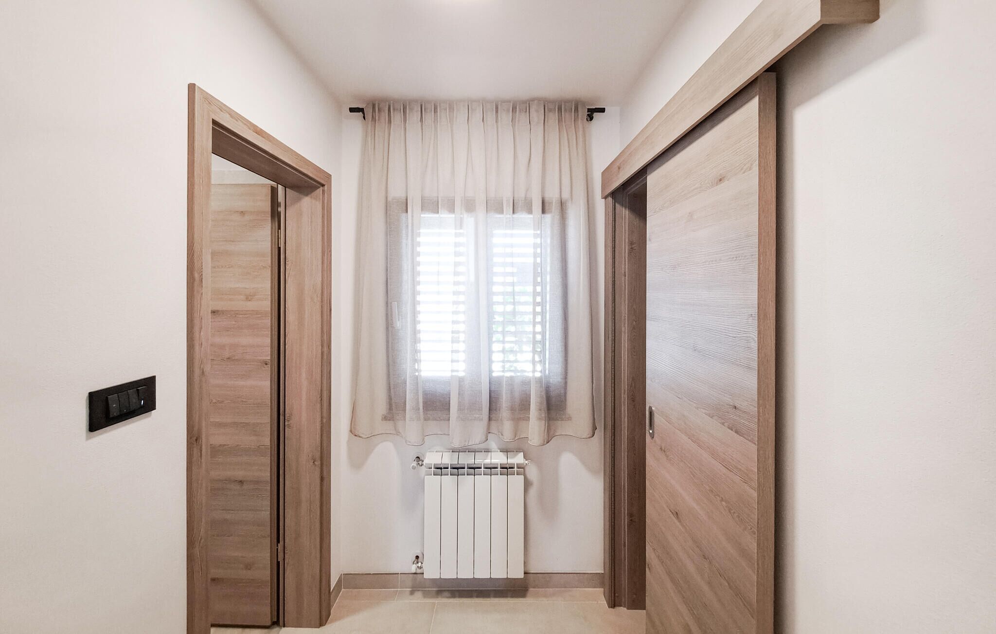Um corredor estreito com portas de madeira e uma janela com cortinas leves.