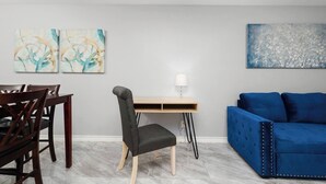 A modern living room with a dining area, featuring a blue sofa, a dining table with chairs, a desk with a lamp, and abstract wall art. - The 3 Queen Modern by 802 & Paredes (Brownsville)