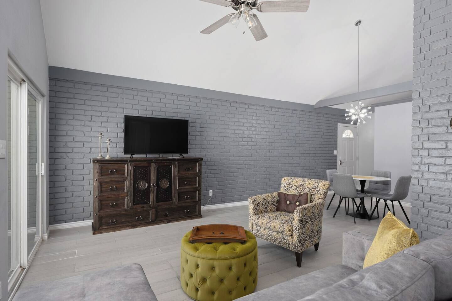A modern living room with a grey brick wall, a wooden cabinet, a television, a patterned armchair, a green ottoman, and a grey sofa.