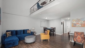 Ein geräumiger Wohnbereich mit einem blauen Sofa, einem runden Hocker, einem gelben Sessel und einem Esstisch mit Stühlen.
