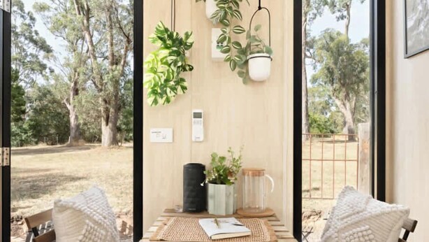 A room with a wooden table, two chairs, a hanging plant, and a view of trees outside. - Delightfully Tiny by Tiny Away (Springmount)