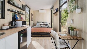 A compact, modern kitchen with a microwave, coffee maker, and storage containers. A small dining area with a table and chair is visible, and a bed with pillows is in the background. - Delightfully Tiny by Tiny Away (Springmount)