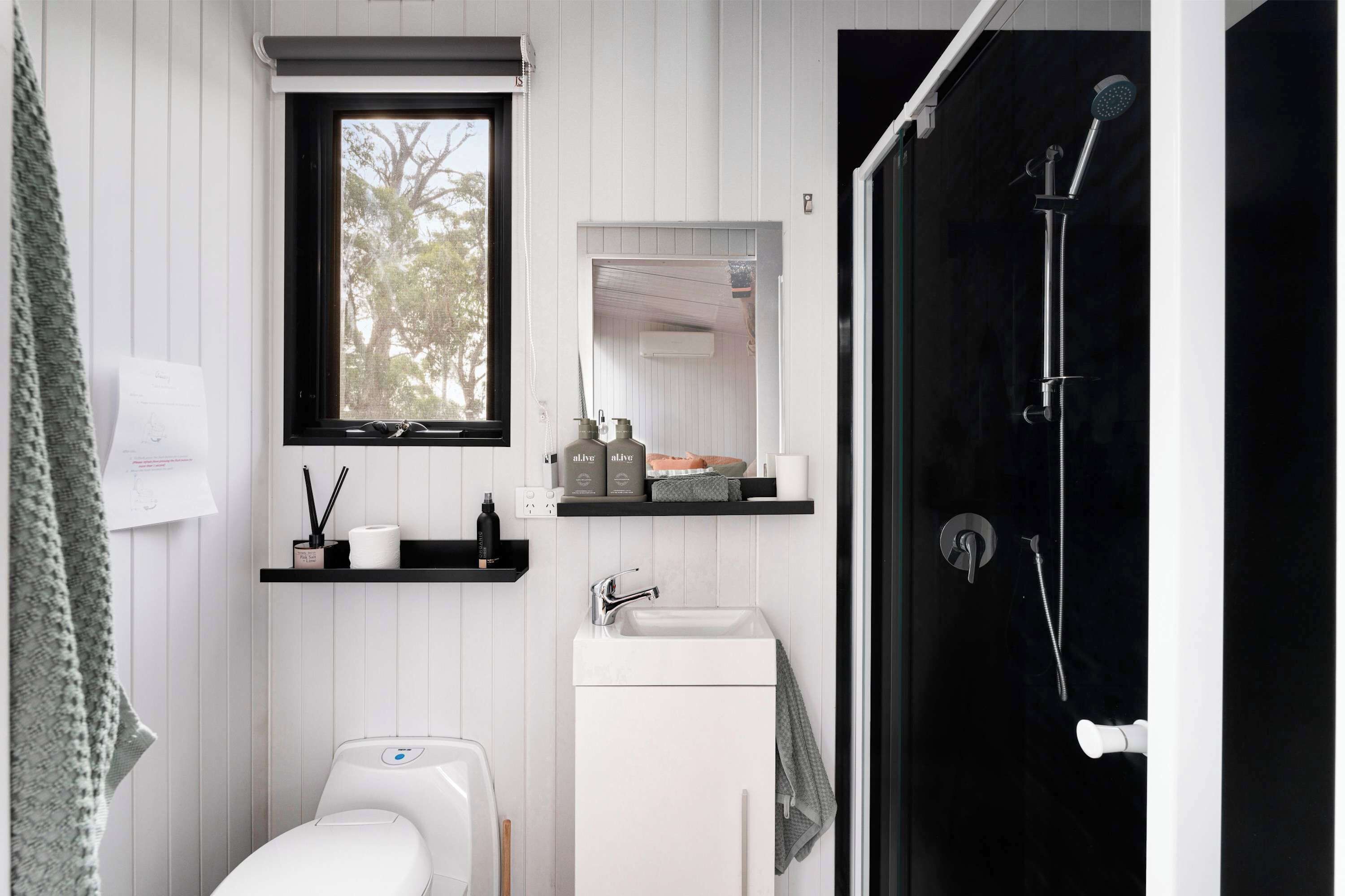 A modern bathroom with a toilet, sink, and shower. There is a window above the toilet, a shelf with toiletries, and a mirror above the sink.