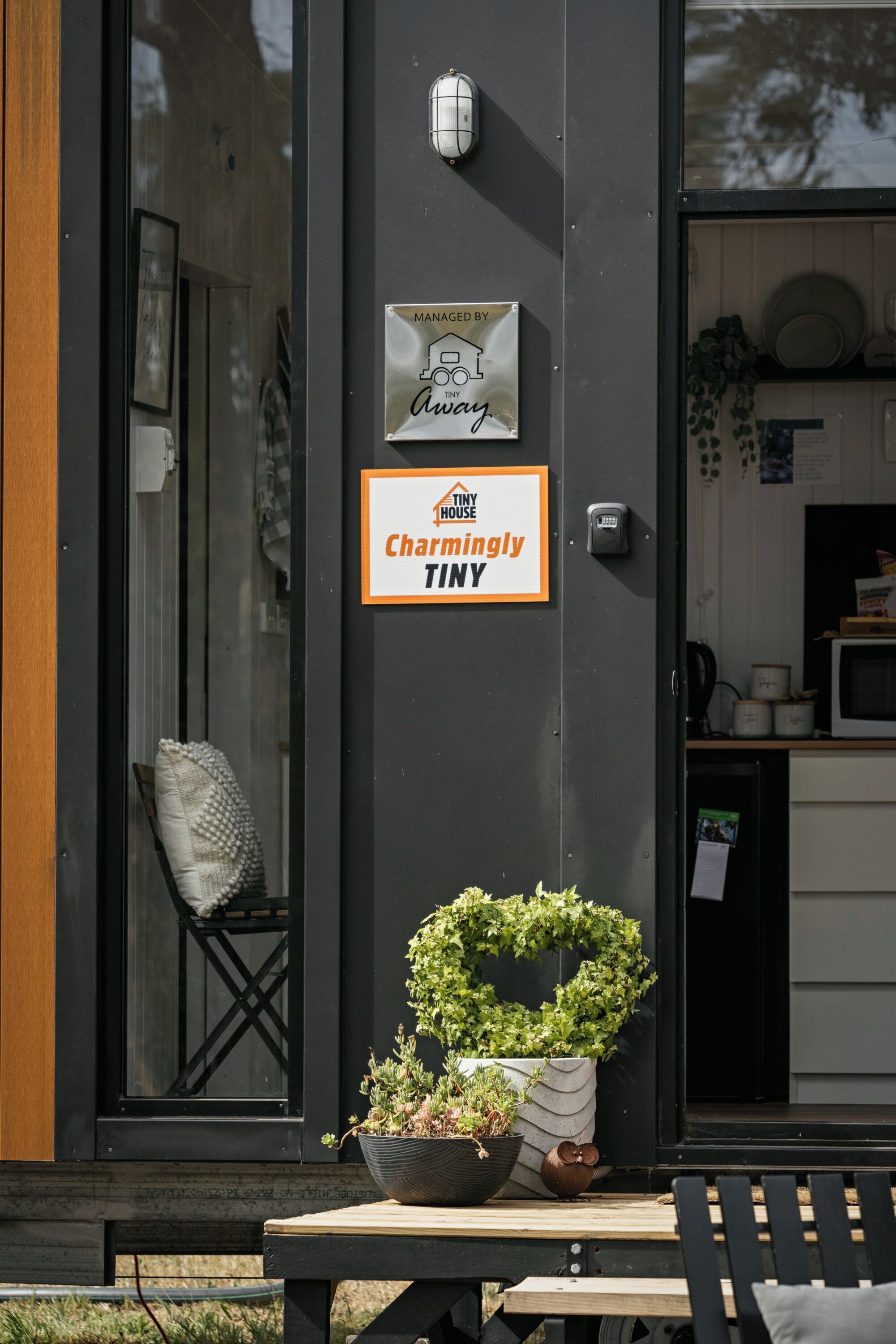A modern building exterior with a sign indicating it's managed by Annie. There's a small window with a cushion, a potted plant, and a wooden table with a bench.