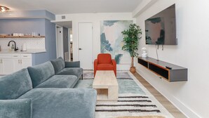 Un salon moderne avec un canapé gris, une table basse en bois, un fauteuil rouge, un téléviseur fixé au mur et un coin cuisine doté de meubles blancs et d’un plan de travail.