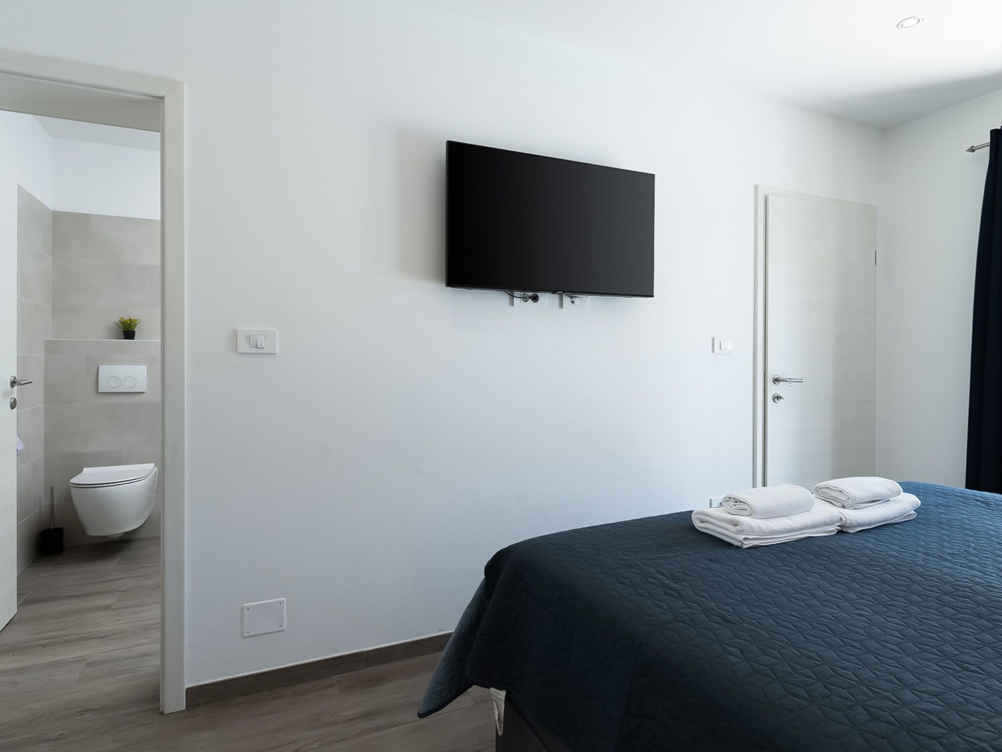 A modern hotel room with a flat-screen TV mounted on the wall, a neatly made bed with folded towels, and a visible bathroom with a toilet and a small plant.