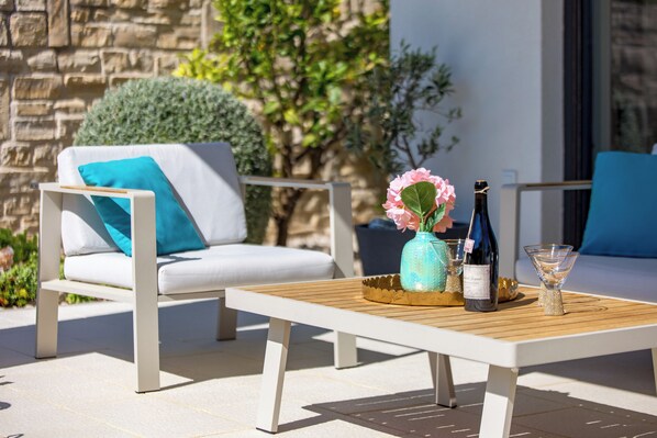 Un área de estar al aire libre con una mesa de madera, dos sillas, un jarrón con flores, una botella de vino y una copa.