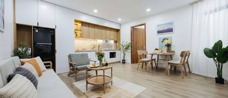 A modern living room with a sofa, chairs, a coffee table, and a dining area with a table and chairs. There is a kitchen area with wooden cabinets and a refrigerator.