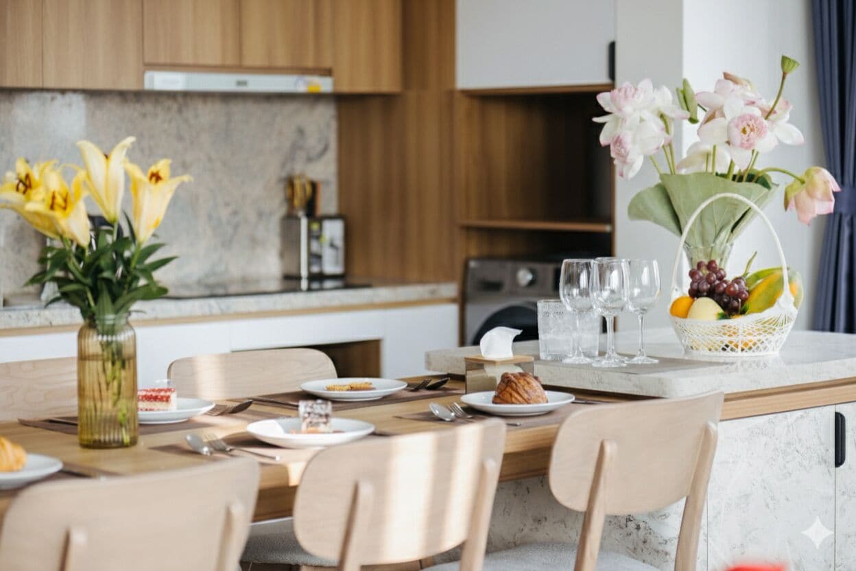 Une cuisine moderne avec des meubles en bois, un plan de travail en marbre et un coin repas avec une table dressée pour deux personnes. On peut y voir des fleurs dans un vase et un panier de fruits sur le plan de travail.