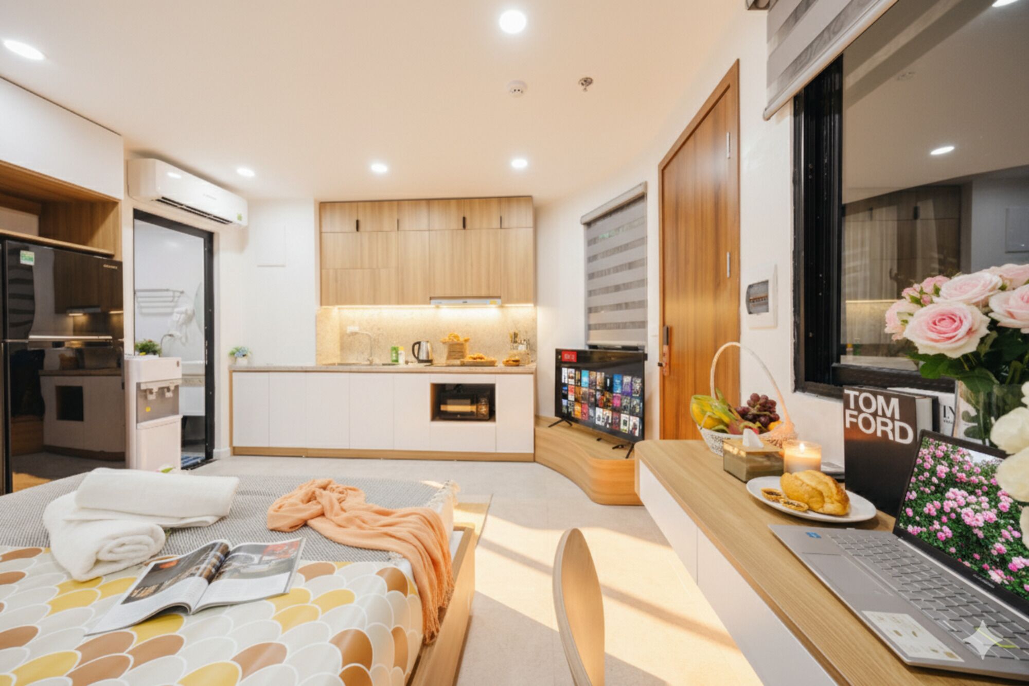 Habitación de hotel moderna con kitchenette, un escritorio con una computadora portátil, una cama con una revista y un florero con flores.