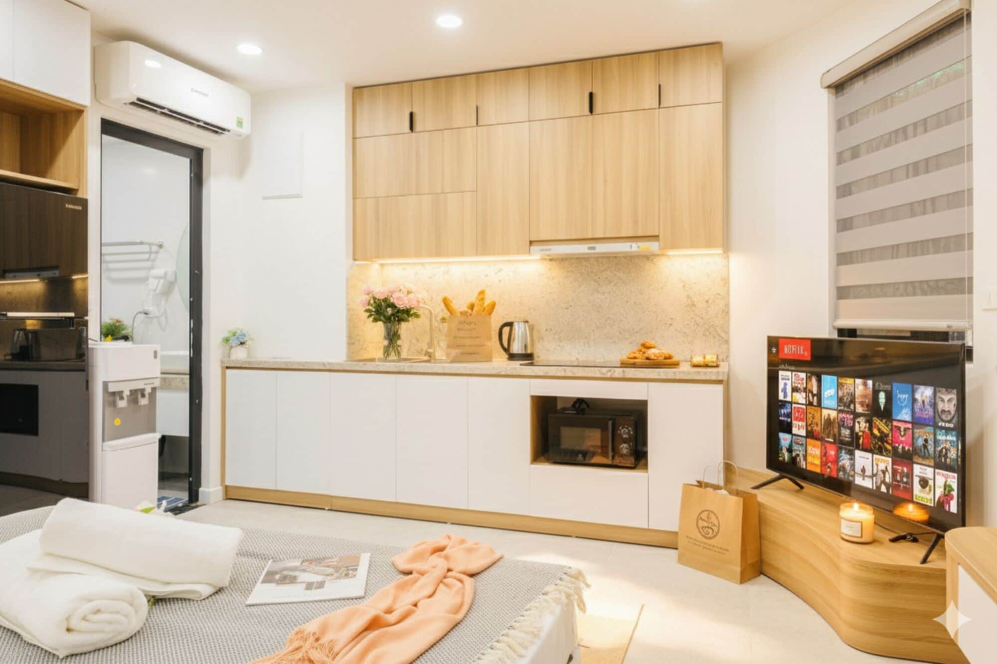 A modern kitchen with wooden cabinets, a built-in oven, and a coffee machine. There is a small fridge with a digital display, a sink, and a countertop with a vase of flowers and a bag of bread.