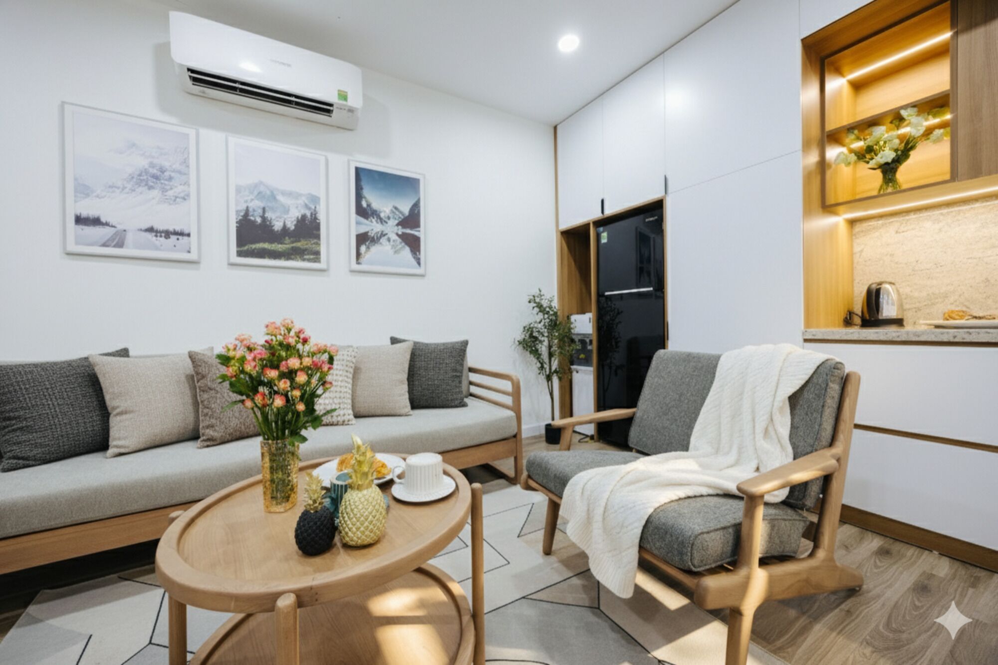 Una sala de estar moderna con sofá, sillón y mesa de centro. Hay tres cuadros enmarcados en la pared, un refrigerador y una ventana con una planta.