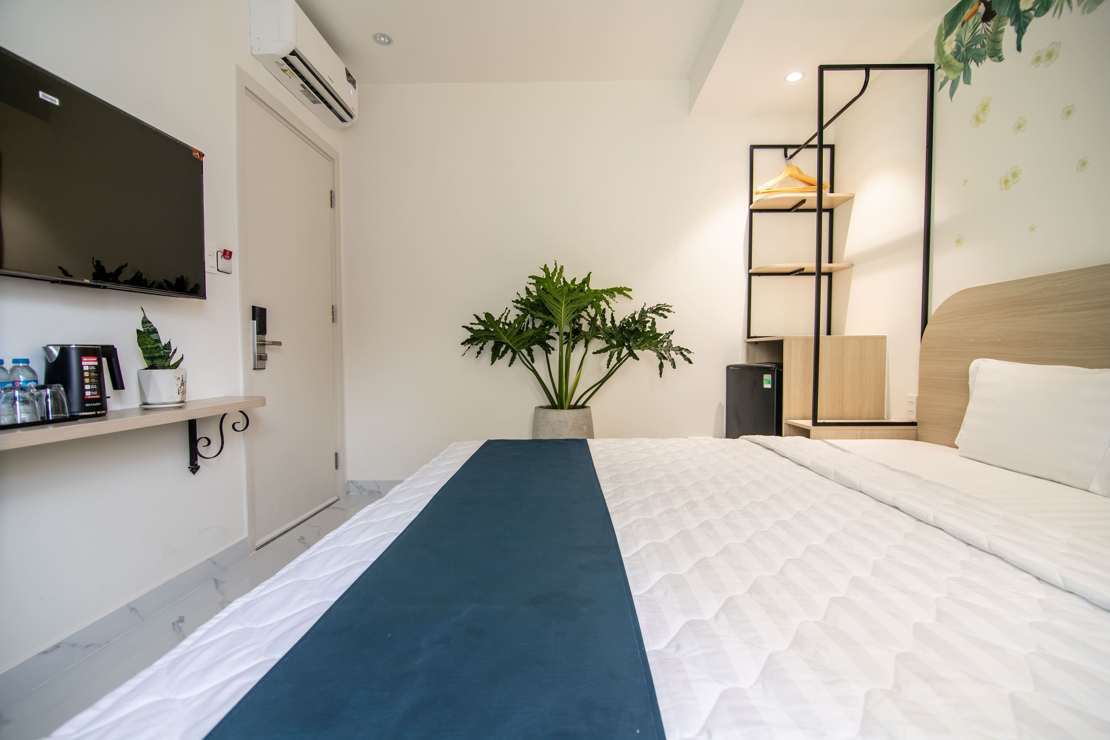 A hotel room with a bed, a TV, a shelf with toiletries, a plant, and a wooden headboard.