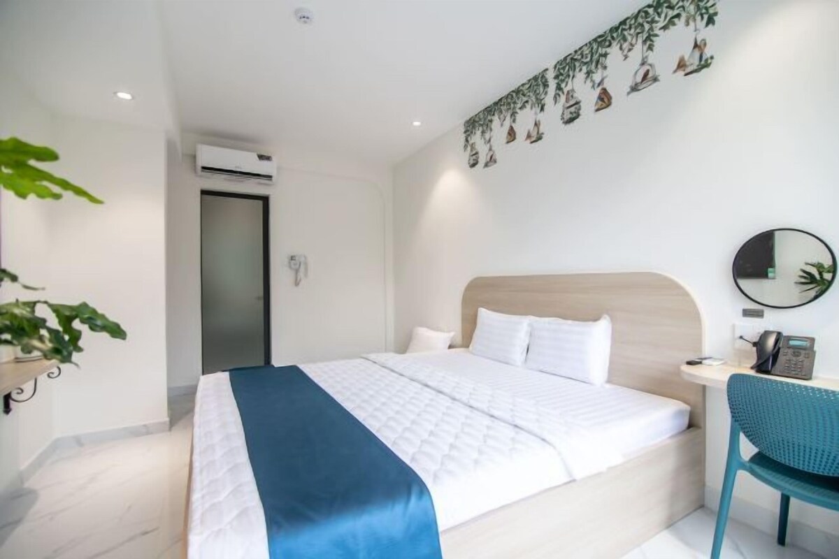 A modern hotel room with a large bed, a desk with a chair, a mirror, and an air conditioning unit.