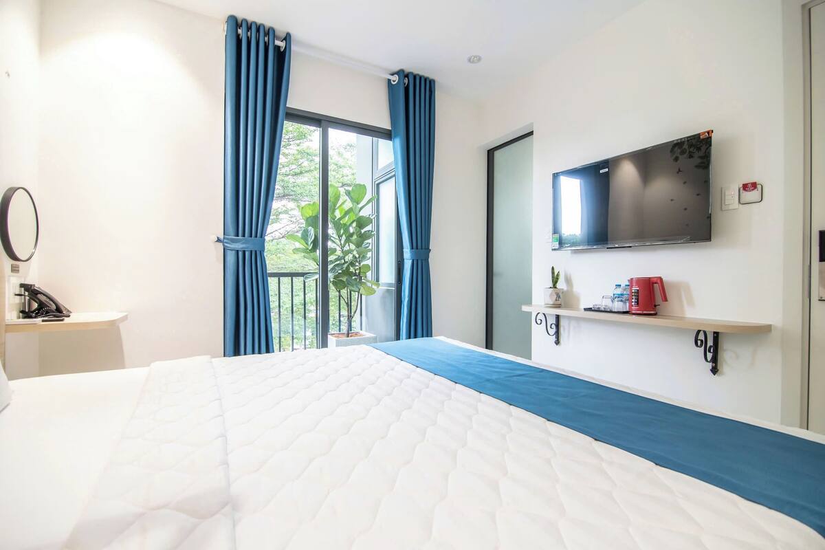 Ein Hotelzimmer mit einem großen Bett, einem an der Wand befestigten Fernseher, einem kleinen Tisch mit Telefon und Pflanze sowie einem Balkon mit blauen Vorhängen.