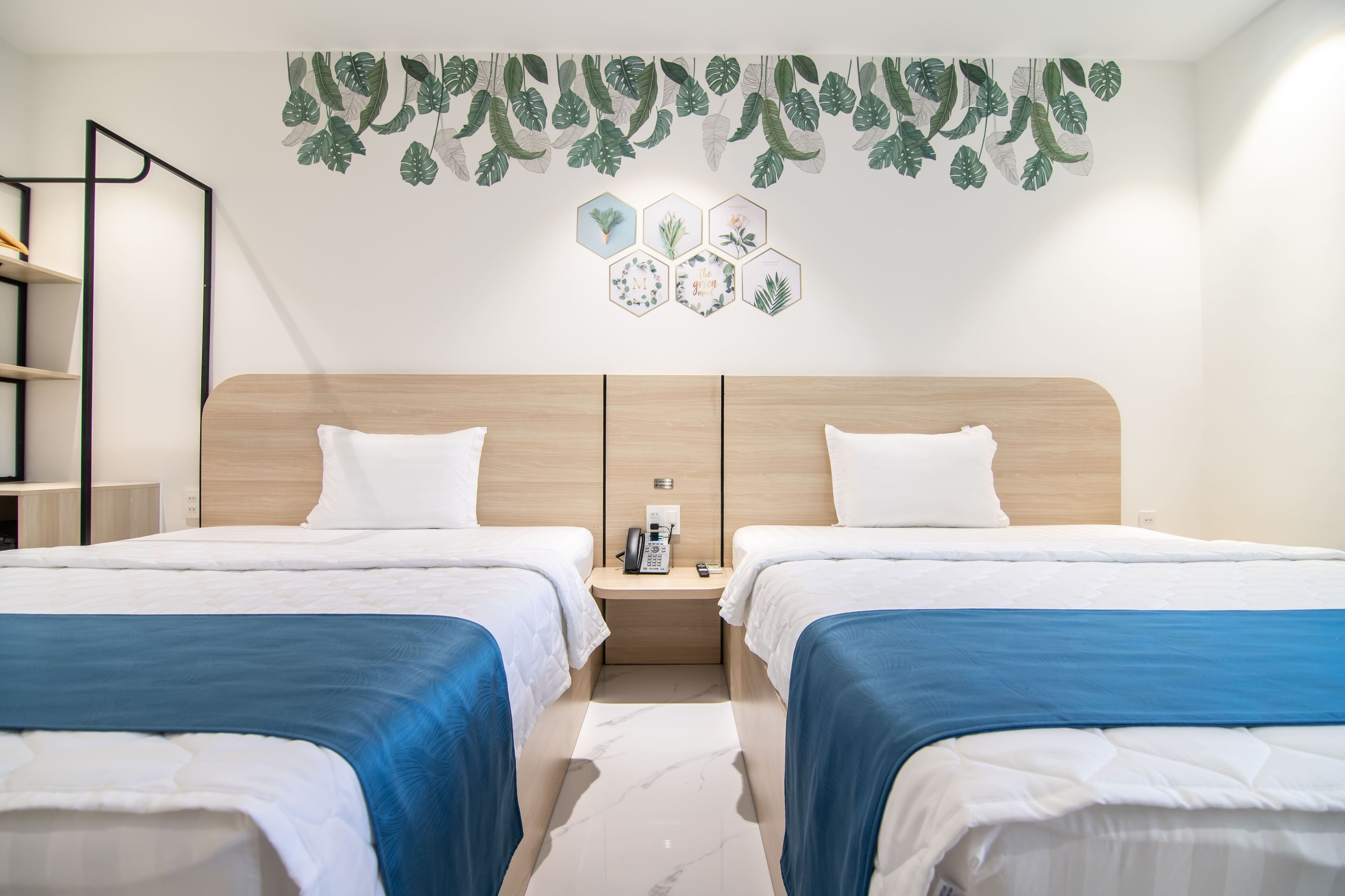 A hotel room with two beds, a wooden headboard, a nightstand with a phone and remote, and a wall with leaf and hexagon decorations.