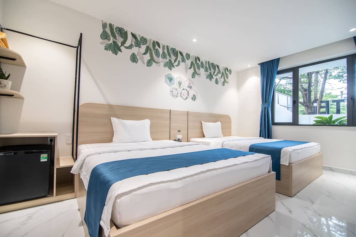 Ein Hotelzimmer mit zwei Betten, einem Schreibtisch, einem Kühlschrank und einem Fenster mit Blick auf Bäume.
