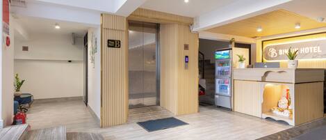 A hotel reception area with a wooden reception desk, a digital screen, and a staircase leading to an upper floor.