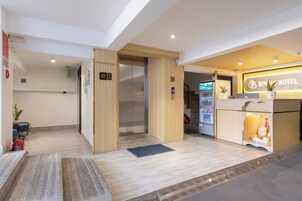 Zona de recepción de un hotel con un mostrador de madera, una pantalla digital y una escalera que conduce a un piso superior.