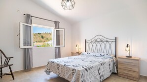 A bedroom with a bed, two bedside tables, a chair, a window with curtains, and a view of a hillside. - Beach front home in Benajarafe (Benajarafe)