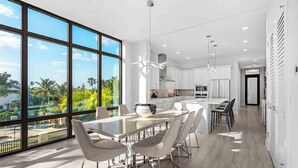 Una cocina moderna con amplios ventanales, un comedor con mesa de vidrio y vistas a palmeras.