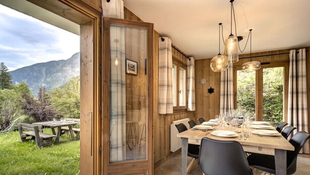 Uma sala de jantar com mesa de madeira posta para seis, uma janela grande com cortinas, e vista para uma paisagem montanhosa.