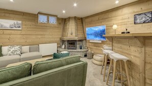 Uma sala de estar acolhedora com lareira de pedra, um sofá, um estofado verde e uma TV fixada na parede.