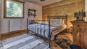 A bedroom with a wooden headboard, a metal bed frame, a window with a view of trees, a radiator, and a small bedside table with a lamp. - Turra - Cosy 80m2 | Central | Remontée | Parking | Calme (Les Houches)