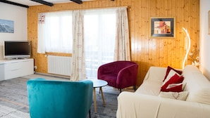 Una habitación con sofá, dos sillones, una mesita, televisor y ventana con cortinas.