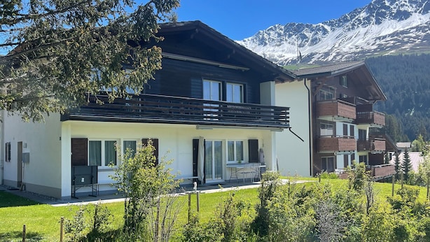 Un moderno chalet de dos pisos con balcón, rodeado de exuberante vegetación y montañas nevadas al fondo.